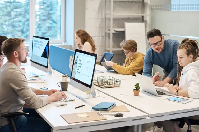 Equipo de trabajo conversa alrededor de computadoras en una oficina iluminada.