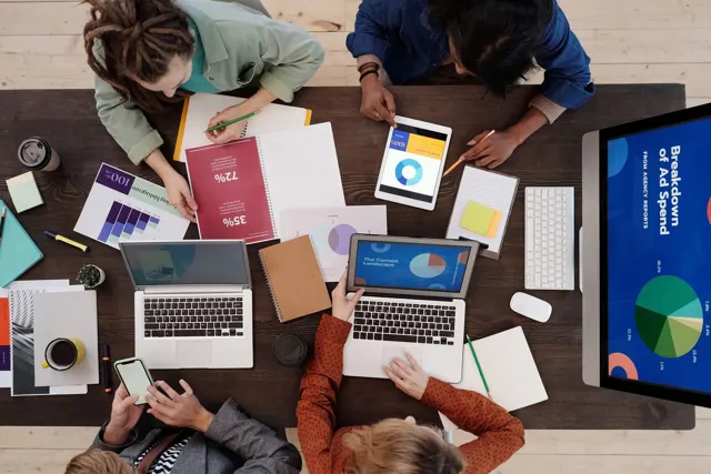 Equipo reunido alrededor de una mesa con computadoras, gráficos y notas de trabajo.